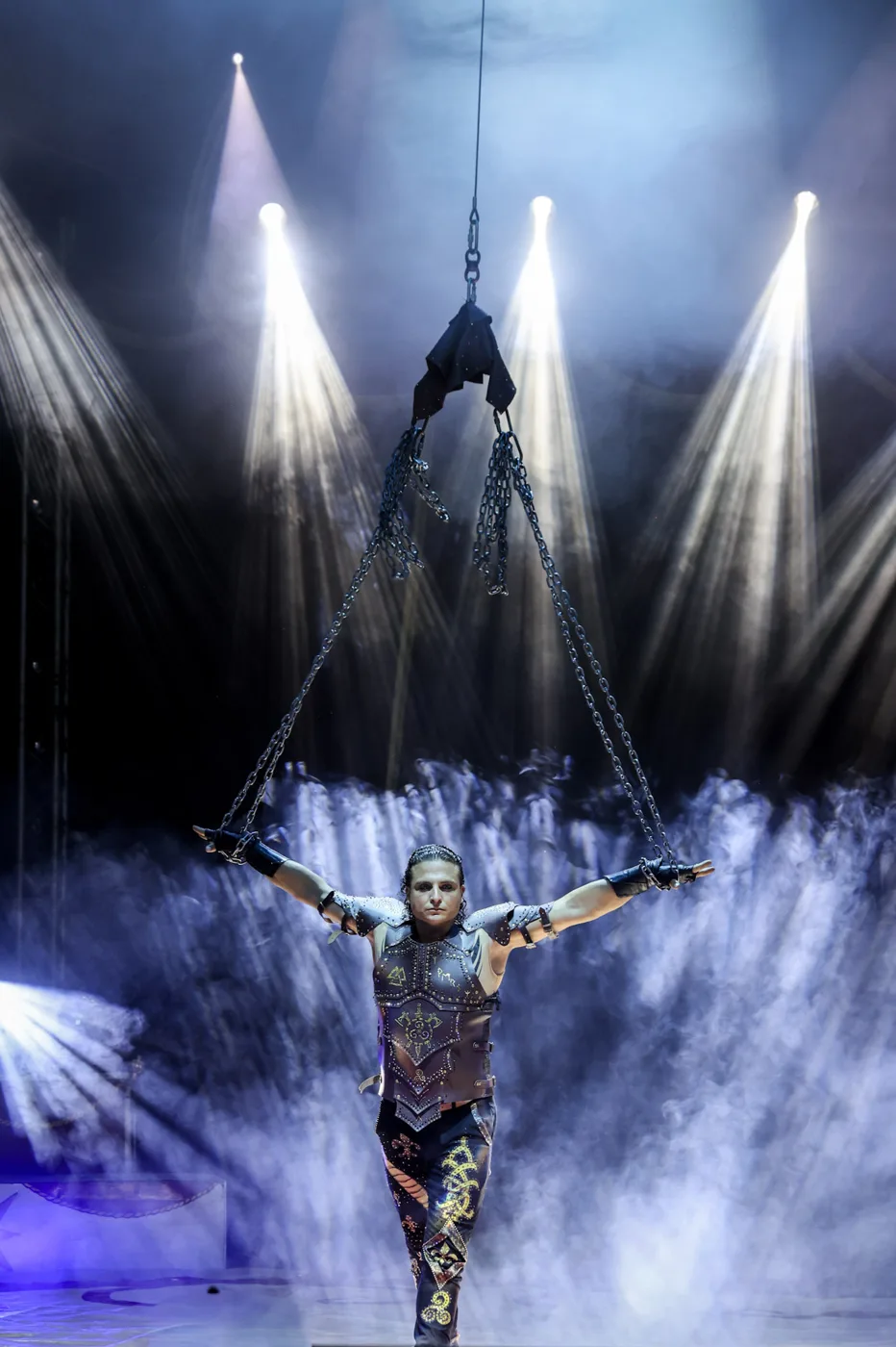 An aerial performer in a studded leather costume lifted on chains through stage smoke and spotlights