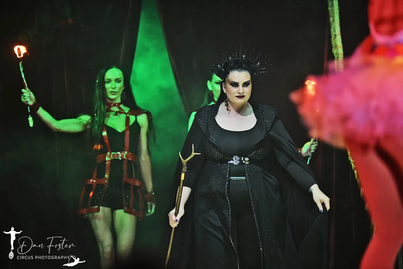 Performers in dark costumes walking through green-lit stage smoke, one in a spiked crown carrying a staff and another holding a flaming torch