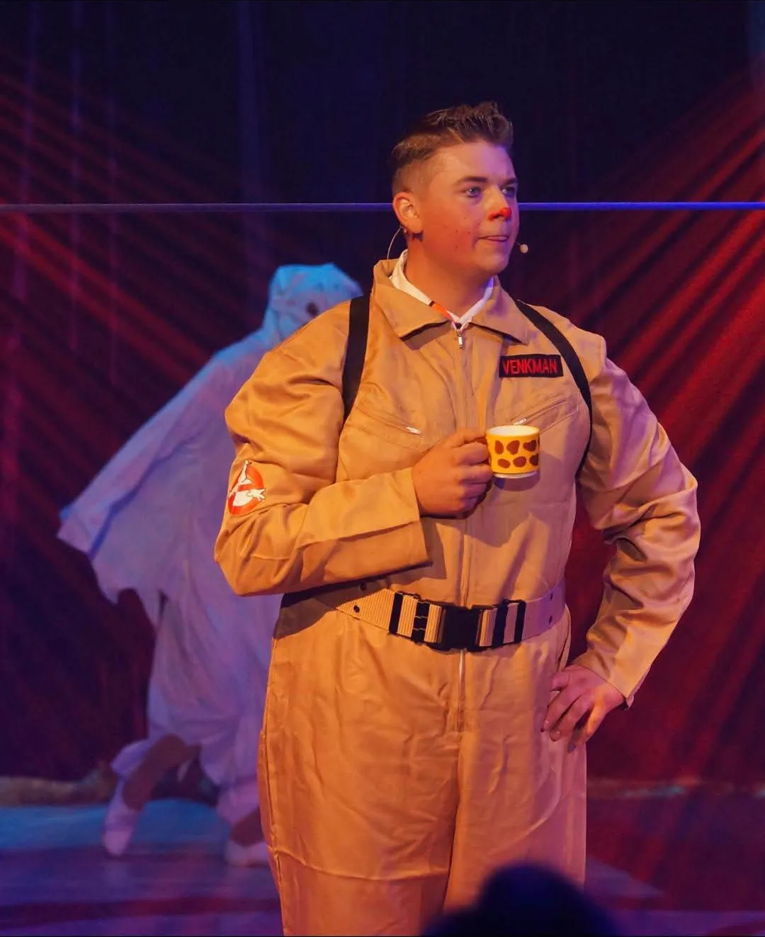 A clown with a red nose in a beige boiler suit holding a small mug, with a performer dressed as a ghost behind