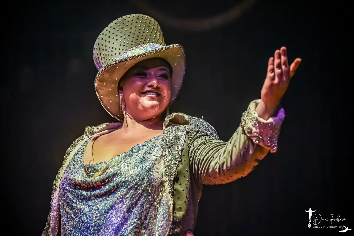 A smiling performer in a sequinned jacket and jewelled top hat, arm raised towards the audience