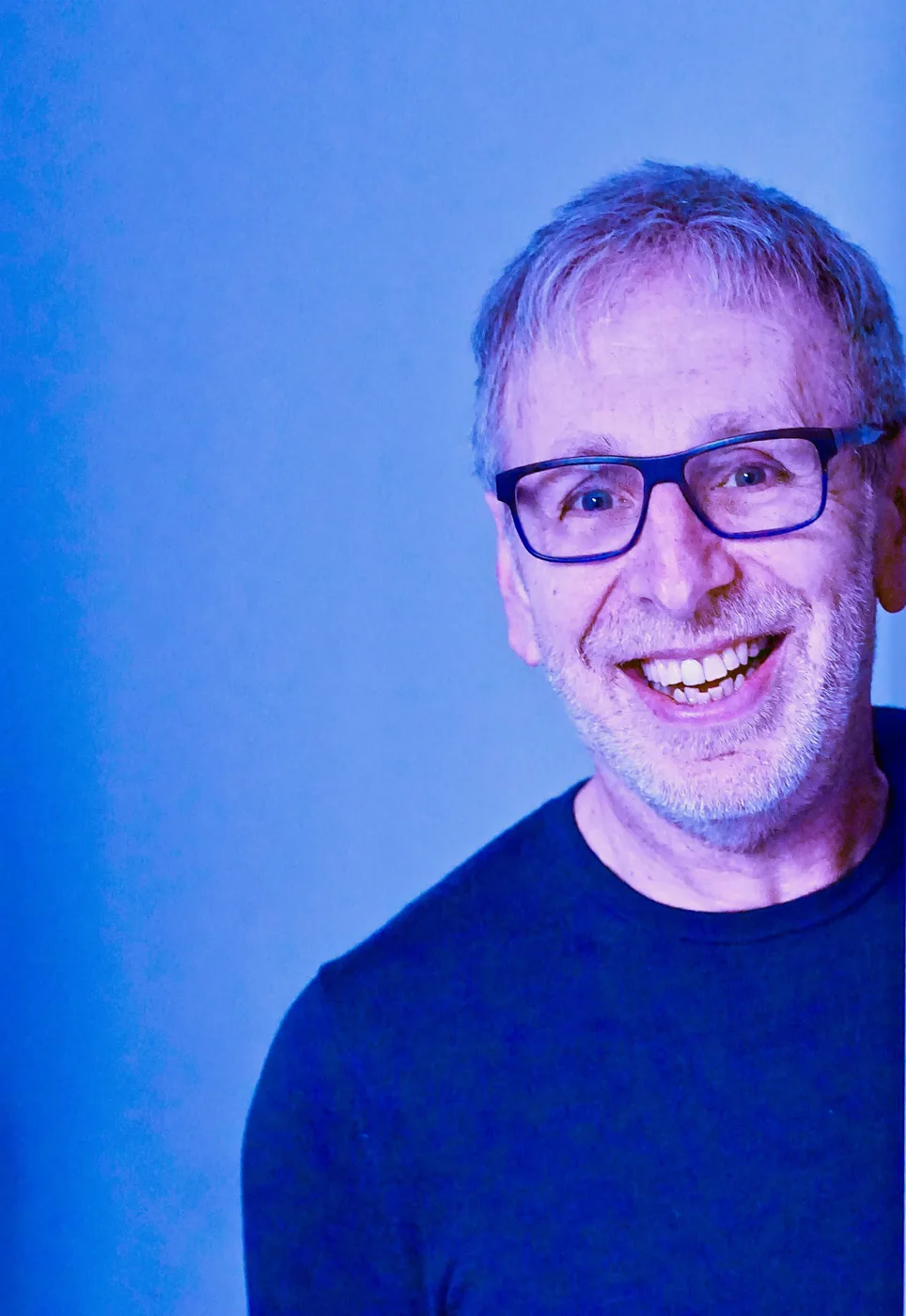 Ian Stone, smiling, in glasses and a dark jumper against a blue background