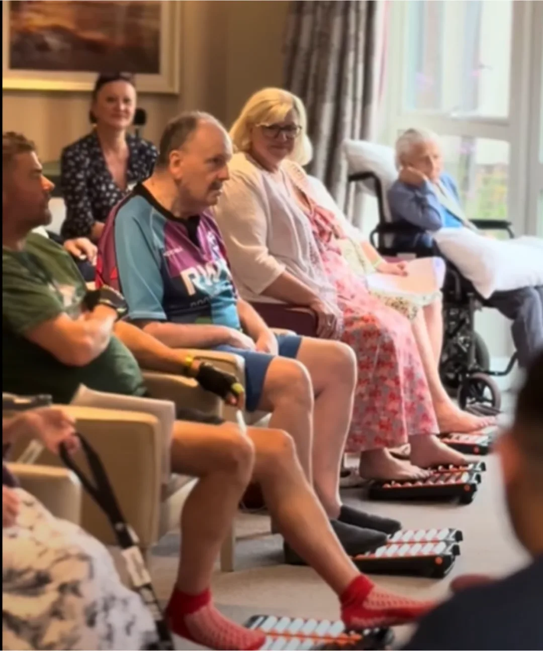 A row of people seated in armchairs and a wheelchair in a care home lounge, each with bare or socked feet resting on a SHUFFLER board