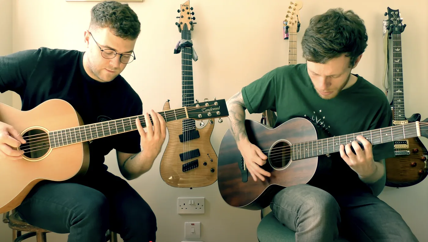 Two acoustic guitars played side by side, with electrics hanging on the wall behind.